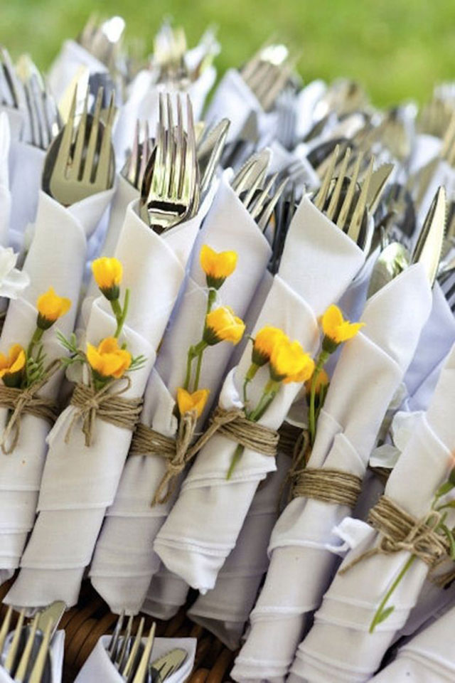 Cubiertos envueltos en servilletas de tela blanca y amarrados con cordel. Como detalle un par de flores silvestres puestas cerca del nudo.