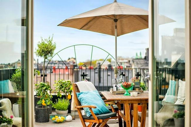 Terraza con mesa y sillas de madera y quitasol. A un costado macetas con limoneros. Sillas con cojines en color celeste.