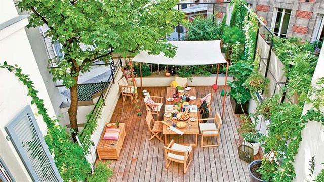 Terraza con una gran mesa y sillas de madera, toldo con techo de lona, banqueta de madera. Enredaderas, arbustos y árboles en macetero por alrededor. Piso de tablones de madera.