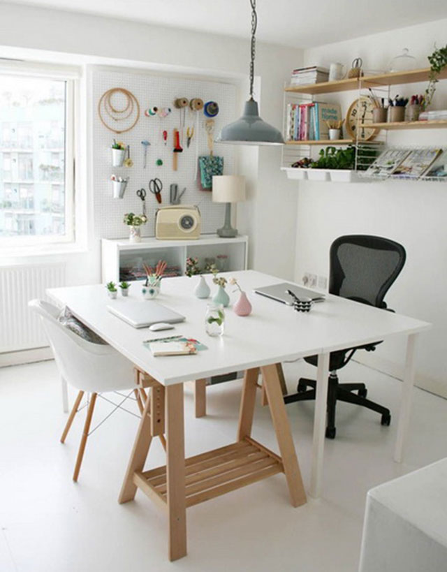 Mesa cuadrada, estructura de madera y cubierta blanca con dos sillas, una blanca y otra negra con ruedas, repisas en los muros, con libros, lápices y plantas. En el muro junto a la ventana un panel con elementos de costura, como bastidores circulares, tijeras y cintas