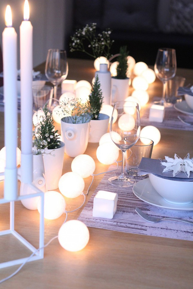 Mesa con vajilla blanca, vasos y copas de vidrio transparente, decorada la mesa con velas blancas largas y una guirnalda de esferas blancas.