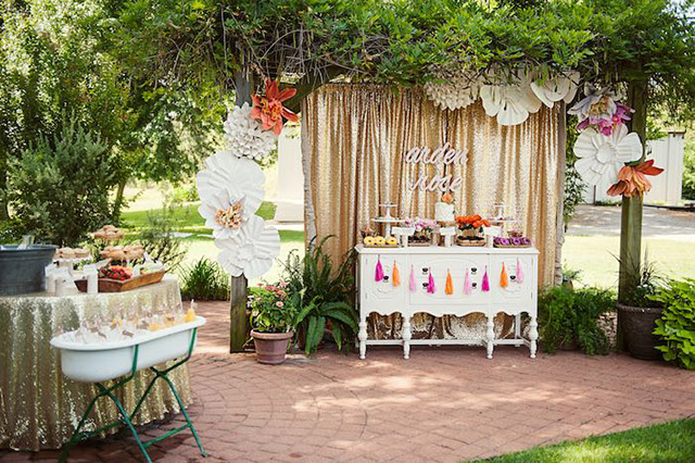 Decoración para una fiesta de cumpleaños de niña en el jardín. Mesa redonda a un costado con mantel dorado. Bajo una pérgola, un mueble estilo vintage blanco con diversos pasteles, organizados tipo buffet, todo enmarcado con flores blancas gigantes.