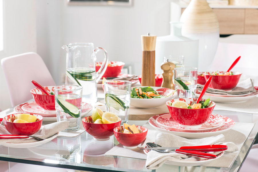 Mesa de comedor con loza de base blanca con diseños en color rojo. Cubiertos tienen base roja haciendo juego. Servilletas color crudo y salero y pimentero de madera.