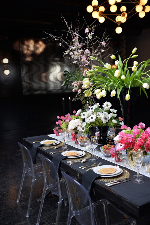 Mesa tipo buffet con textil en negro, sillas de acrílico transparente. Sobre el camino de mesa color blanco varias macetas con flores blancas y rosadas y un gran florero negro con tulipanes amarillos.