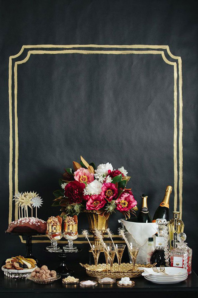 Buffet de año nuevo. Decoración con copitas doradas sobre bandeja también dorada, florero dorado sobre plato de torta negro con flores fucsias y blancas. Sobre el muro de fondo un lienzo negro con un marco dorado dibujado.