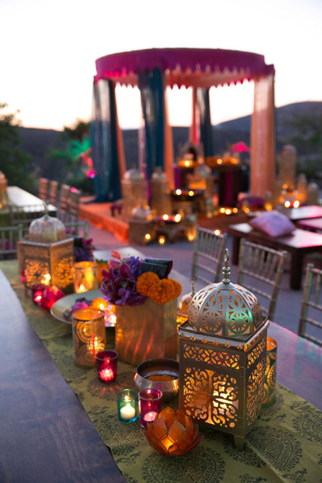 Mesa con caminito de tela cubierta de faroles, porta velas, fanales y flores en florero y fuente, todo en un estilo muy arabesco.