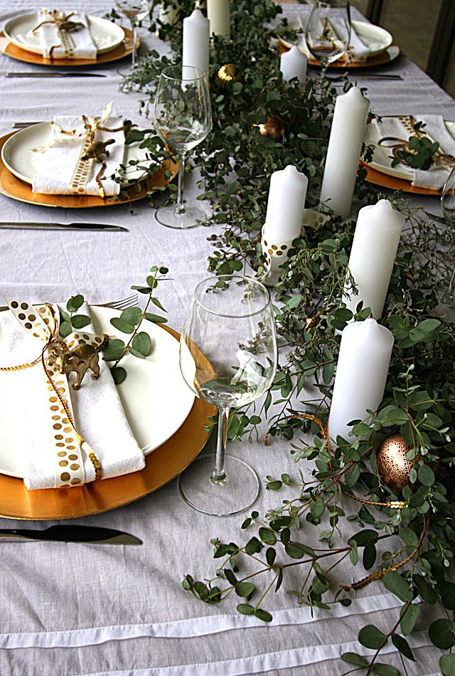 Ramas verdes decorando como camino de mesa y entre medio velones blancos. Los platos dorados también se encuentran decorados con algunas ramitas y sobre el plato unas pequeñas figuras de animales dorados.