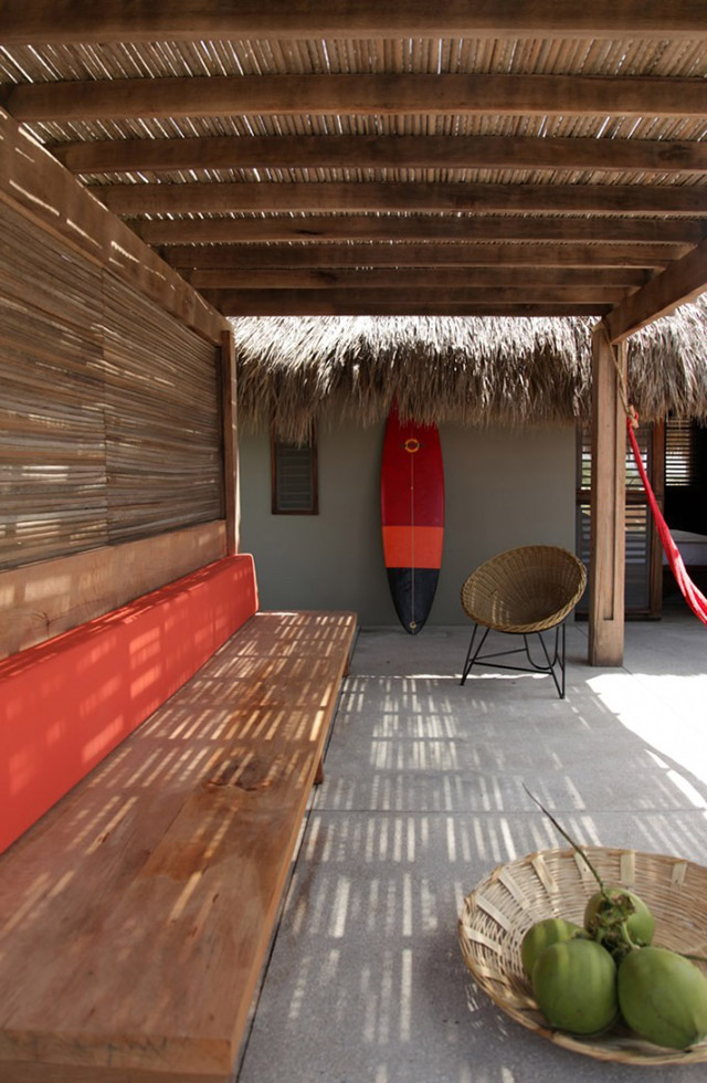 Terraza de casa en la playa con muro y cielo recubiertos de bambú. A lo largo del muro, una banqueta de madera con cojín rojo del largo del respaldo. Al fondo apoyada en la pared una tabla para surf en colores rojo, anaranjado y negro.