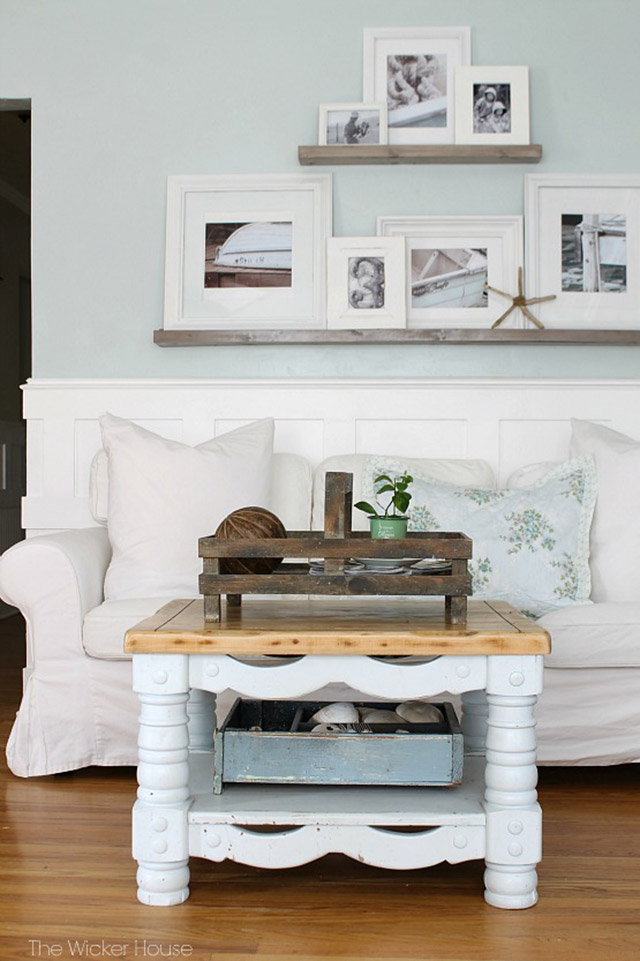 Sala de estar con sofá de tela en color crudo y mesa de centro de madera con la base pintada en blanco y la cubierta en color natural. En el muro del sofá, dos repisas de madera con porta retratos de marco blanco.