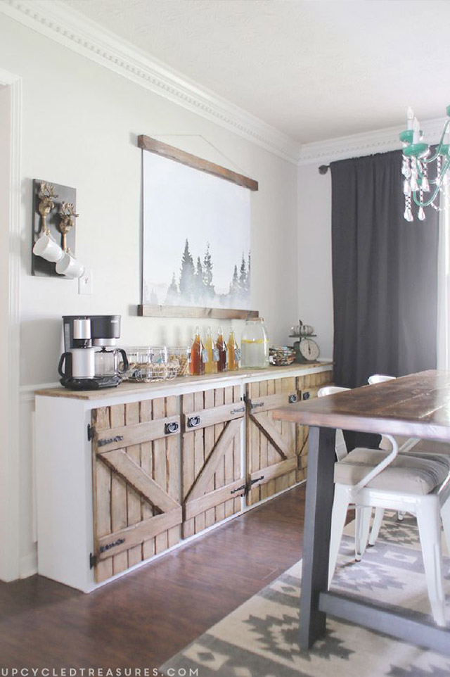 Comedor con gabinete adosado a la pared, blanco con puertas de tablones de madera y bisagras y manillas de fierro. La cubierta del gabinete dispuesto como buffet con cafetera, vasos, botellas de licores y jarrón de jugo. Mesa de comedor de estructura metálica negra y cubierta de madera natural, sillas metálicas blancas con cojines en los asientos.