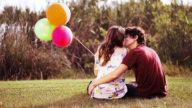 Pareja de espaldas sentadas en el pasto besándose.