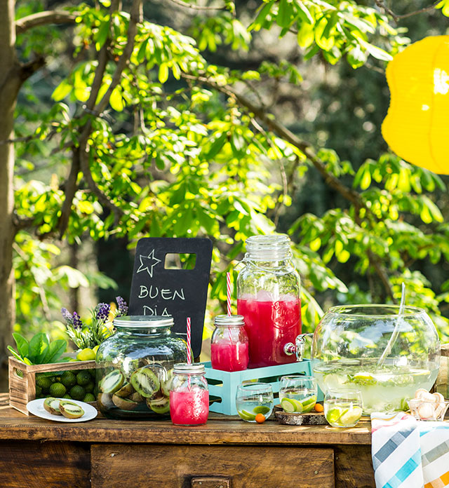 Buffet con dispensadores de líquidos frutales y copas y vasos con diversas frutas.