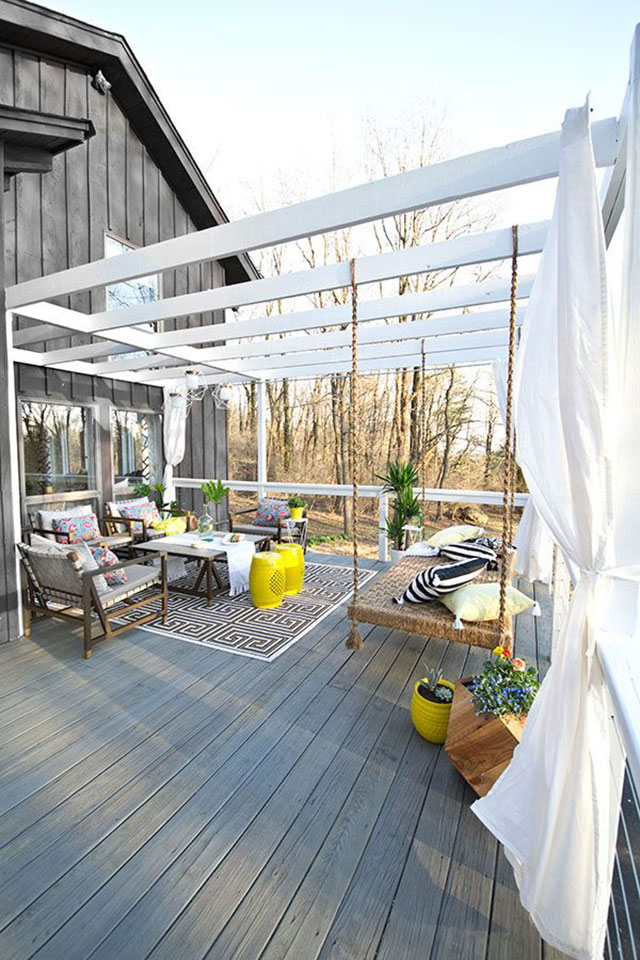 Terraza de piso de madera. Del muro de la casa nace una pérgola de madera pintada blanca. Juego de terraza de madera con cojines grises y multicolores. Alfombra bajo el juego de terraza de diseño geométrico en marrón y blanco, pisos de cerámica color amarillo. Un gran columpio con cojines amarillos y blancos con negro, cuelga de las vigas de la pérgola.