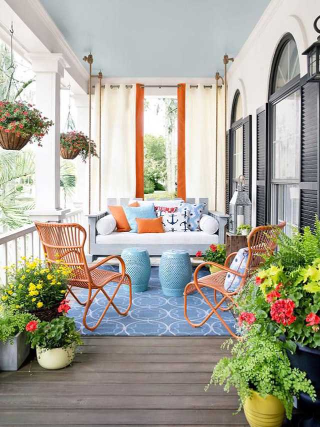 Terraza rodeada de macetas con plantas y flores, dos sillas de mimbre pintadas color anaranjado, dos pisos de cerámica color celeste, sofá de madera con cojín blanco en el asiento y muchos cojines de colores celeste y anaranjados y con motivos náuticos. Macetas colgantes desde el techo y sobre la baranda de la terraza.