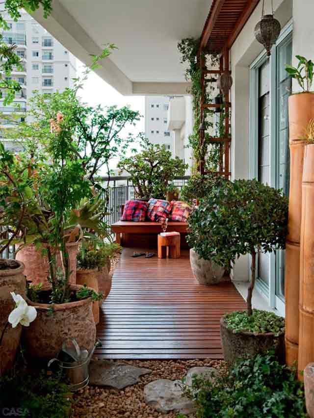 Balcón con el piso recubierto de piedras pequeñas y decking. Sillón de madera con respaldo bajo y cojines. Rodeado de macetas con plantas, enredaderas y árboles pequeños. La decoración es estilo oriental con bambú y lámparas colgantes.