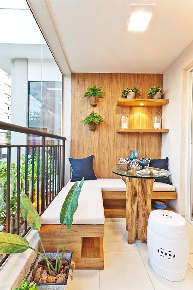 Balcón con piso cerámico, banqueta esquinera de madera y cojín blanco en el asiento, mesa de centro con tronco de base y cubierta de vidrio. Las plantas decoran el espacio con una jardinera de latón a un costado de la banqueta y maceteros sobre el muro y en repisas. Cojines azules sobre la banqueta y piso de cerámica blanco.