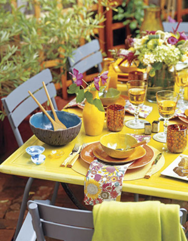 Detalle de mesa de patas metálicas y superficie de madera pintada amarilla. Menaje en colores grises, azules, anaranjados y amarillos, de loza con bordes irregulares, servilletas de tela y estampado de flores.