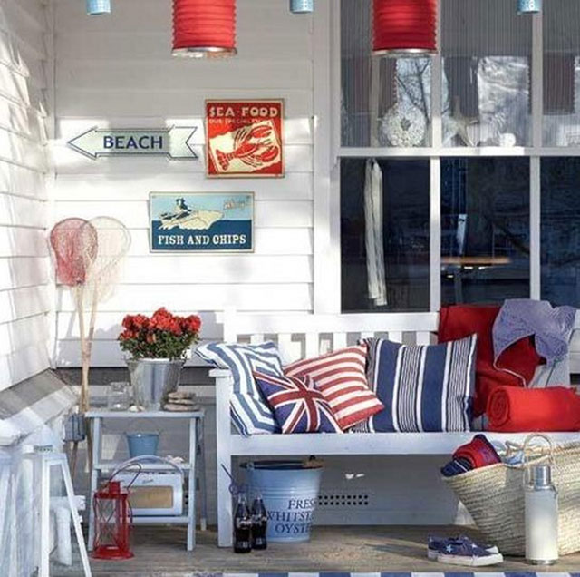 Sillón de madera blanco  junto al ventanal en la terraza. De la pared de madera cuelgan placas metálicas con señaletica marina y anuncios de comida. Junto al sillón, que se encuentrta cubierto de cojínes en colores azul, blanco y rojo en estilo marino, una mesita de arrimo con un balde metálico con flores rojas.