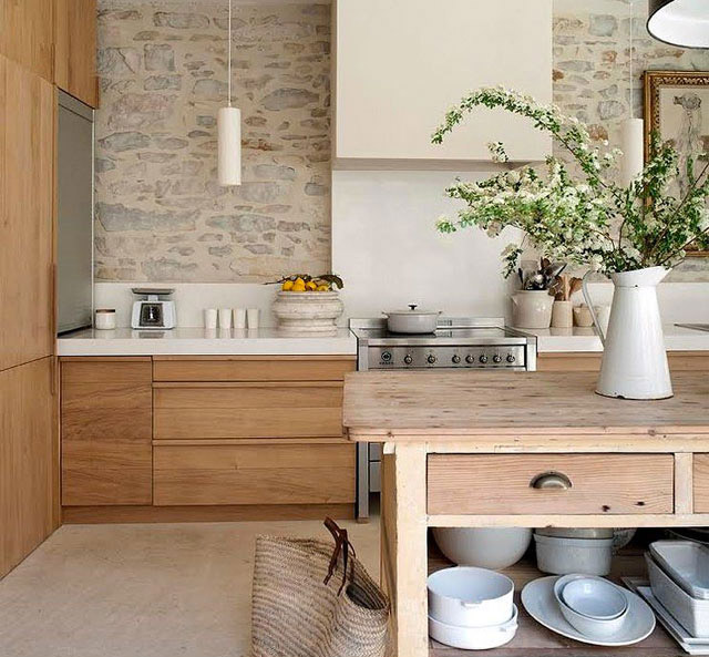 Cocina con muebles de madera y encimera blanca. Al centro se ve un mesón con cajones de madera color natural, en la parte inferior el menaje en tonos blancos. Sobre el mesón un jarrón enlozado blanco con ramas en flor.