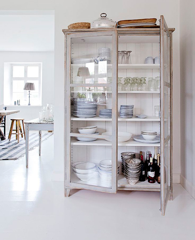 Vitrina de madera blanca desgastada con dos puertas con vajilla, vasos y botellas en el interior