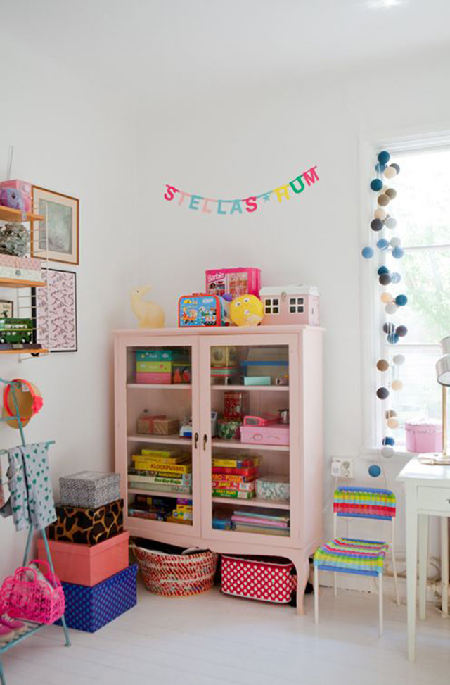 Dormitorio infantil con una vitrina rosada de dos puertas con patas. En el interior de la vitrina de cuatro niveles cajas de distintos colores y, bajo las patas, un par de canastos