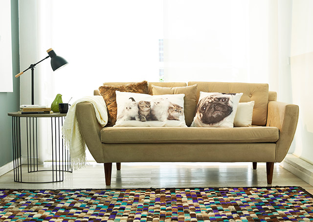 Sofá beige con patas de madera con cojines estampados de animales, alfombra multicolor, cortinas blancas. Homy