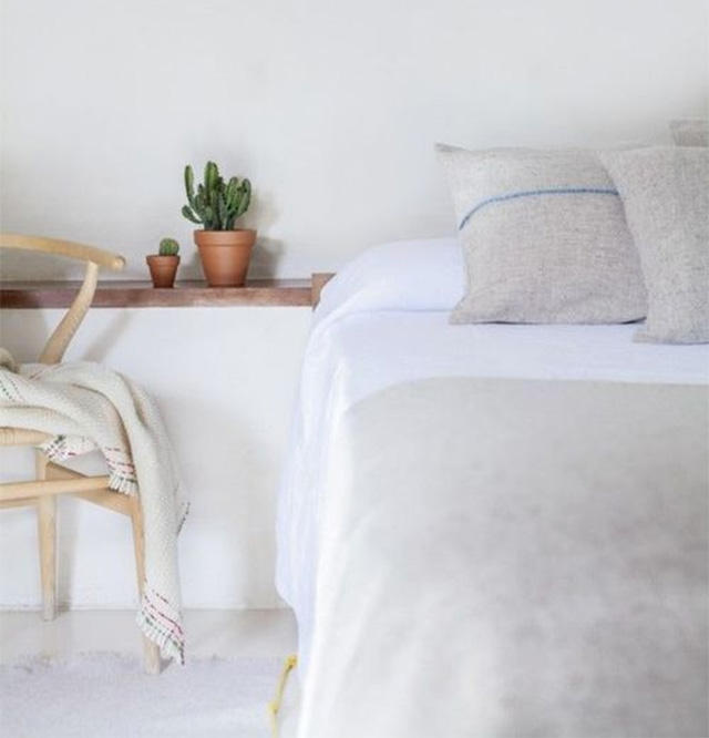 Cama en dormitorio minimalista, sábanas blancas, cobertor gris con cojines a tono, velador tipo repisa con dos maceteros con cactus