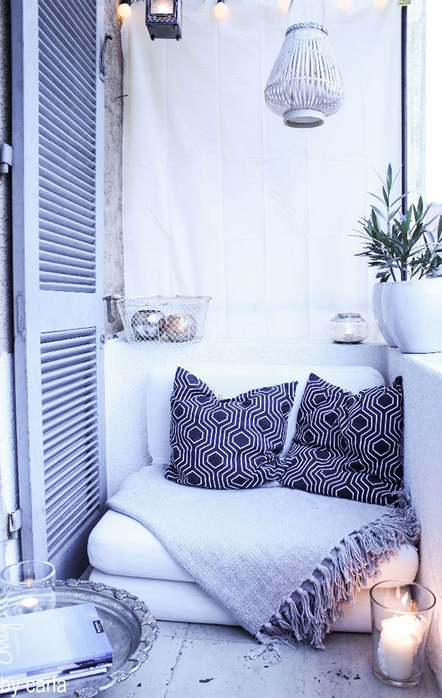 Balcón estilo mediterráneo. Un sillón blanco con manta gris y dos cojines con diseño en azul y figuras geométricas blancas. En los bordes del balcón un par de maceteros blancos y una canasta con porta velas. Del techo cuelgan dos faroles.