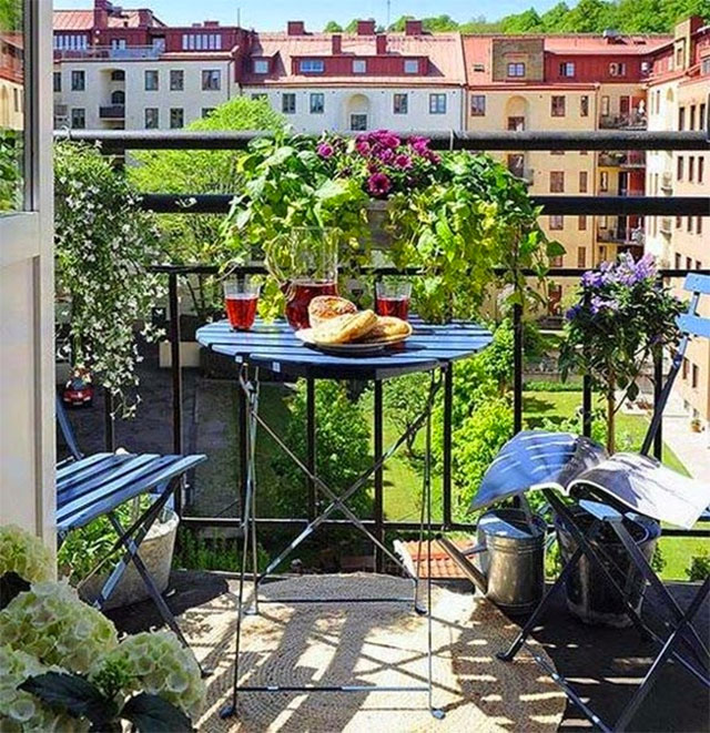 Juego de terraza de dos sillas y mesa plegables de color verde oscuro. La mesa está servida con una jarra de jugo y dos vasos más un plato con pan. De la baranda del balcón cuelgan maceteros con plantas y flores.