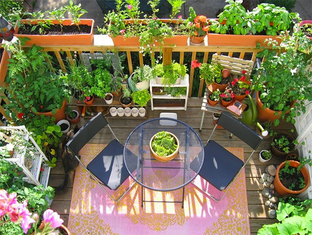 Balcón desde arriba, podemos ver un juego simple de terraza, una mesa redonda y dos sillas sobre una alfombra de diseño en amarillo y rosado. Todo rodeado de maceteros y jardineras con pequeños árboles, plantas aromáticas y flores, algunas jardineras sirven de huerto.