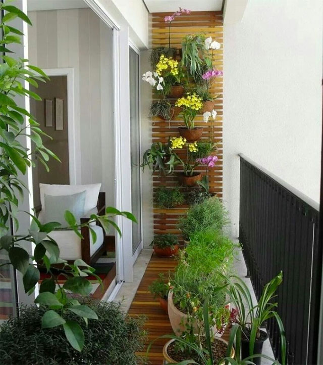 Balcón utilizado como un pequeño jardín con un panel de listones de madera del que cuelgan maceteros con plantas, suculentas y flores. En el piso siguiendo la linea delpanel, un caminito de madera y sobre el más maceteros con plantas.