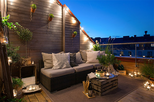 Terraza con sillón de ratán y mesita de centro de pallet iluminada con fanales de vidrio ubicados alrededor de los muebles de terraza.