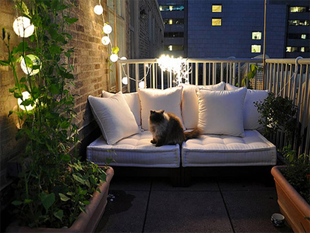 Balcón con un sofá de dos cuerpos de cojines blancos y un gato encima, jardineras con plantas trepadoras. Iluminación de guirnalda de ampolletas de luz blanca.