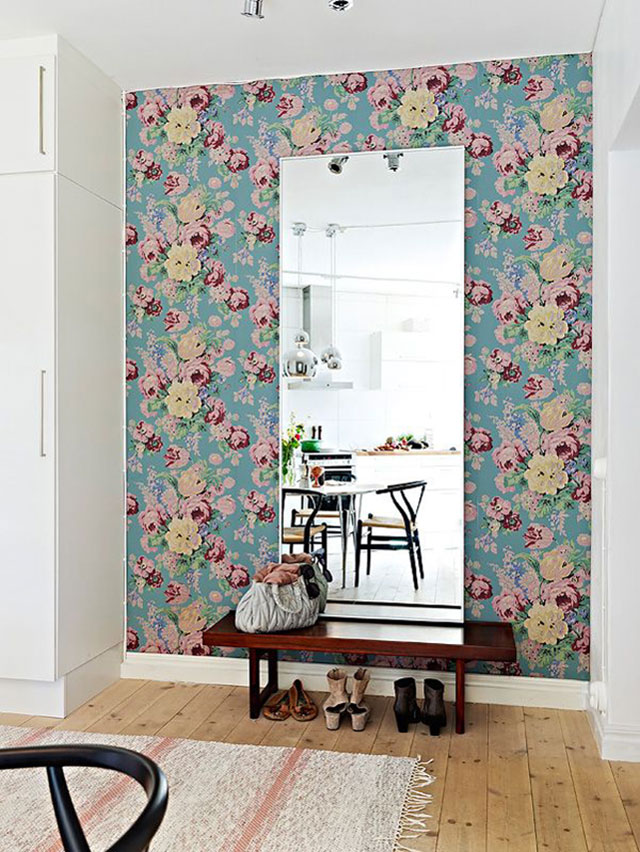 Vista de un muro de un hall de entrada. Papel mural floreado fondo turquesa y flores rosadas y amarillas, un espejo, una banqueta baja con una cartera encima y debajo un par de zapatos.