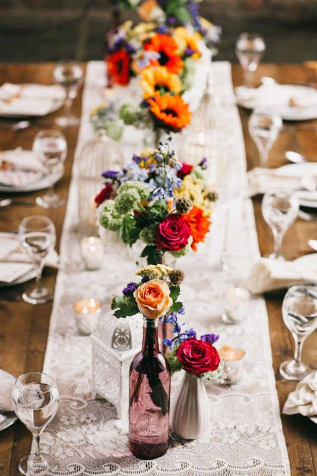 Mesa decorada con un caminito de mesa, velitas y varias botellas con ramos de flores multicolor.