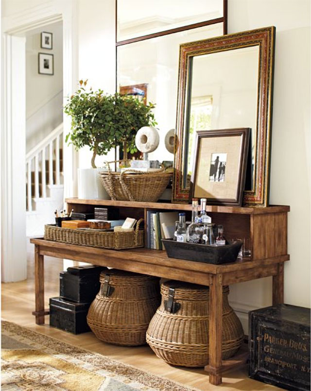 Entrada estilo rústico con mueble tipo arrimo de madera, decoración con canastos de mimbre sobre el mueble y canastos más grandes bajo el mueble. Espejo y marco con foto encima del mueble.