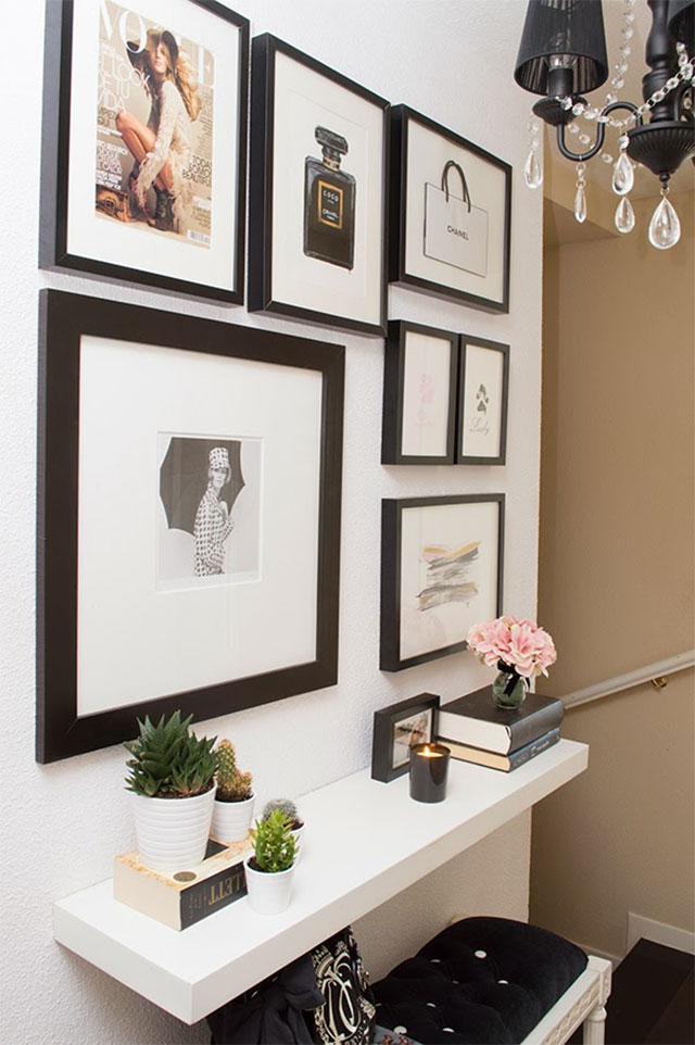 Mural con fotografías e ilustraciones en marcos negros. Repisa blanca dispuesta como arrimo, encima un par de cactus, libros, una vela y un pequeño florero con una flor grande. Bajo la repisa una banqueta acolchada en tela negra y madera blanca.