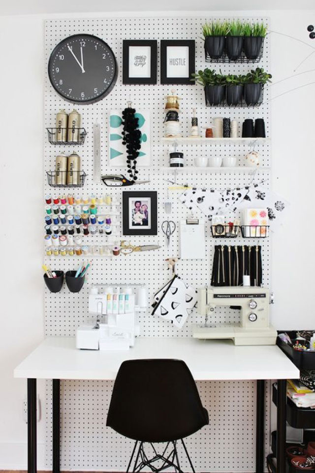 Mesa de escritorio de superficie blanca y patas negras, silla negra. Sobre la mesa dos máquinas de coser. Muro con panel de madera perforado blanco con organizadores para los hilos, las tijeras, un reloj, un par de maceteros con plantas y fotografías e ilustracioneas enmarcadas.