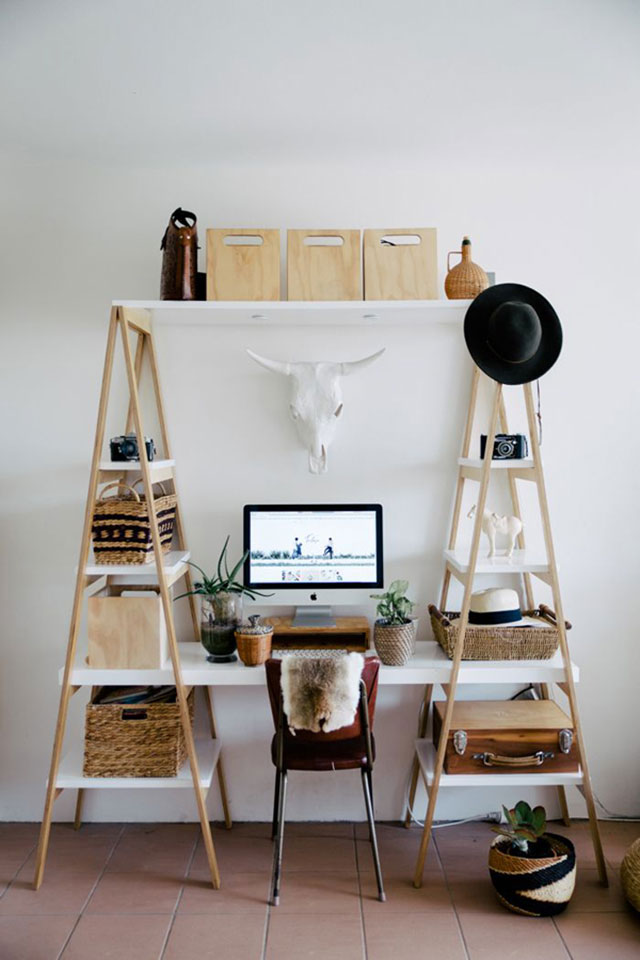 Escritorio armado con un par de estantes tipo escalera de madera. La mesa de escritorio es un tablón de madera blanca soportada por los estantes. En la mesa un monitor sobre una caja de madera para guardar el teclado, una suculenta y una planta. Los estantes con cajas de madera y canastos de mimbre.