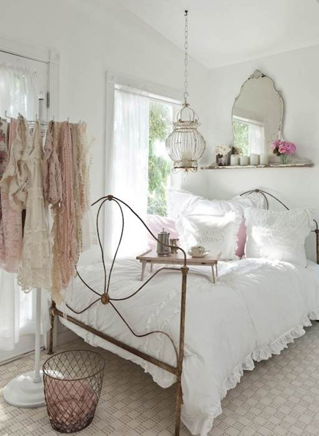 Dormitorio femenino en tonos blancos. Cama de estructura metálica y cubrecama blanco y almohadones blancos y rosados. Espejo con repisa en la pared sobre la cabecera de la cama, en la repisa un par de velas blancas y floreros con flores rosadas y blancas. Desde el techo y sobre la cama una lámpara jaula estilo vintage.