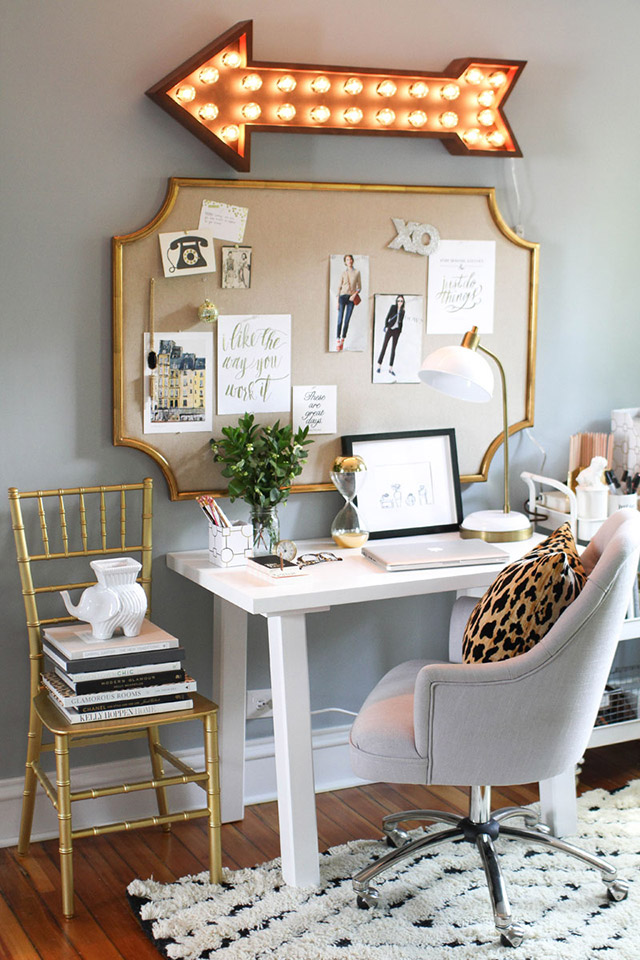 Escritorio con una mesa simple de madera blanca, silla de escritorio tipo poltrona, una silla dorada al costado de la mesa con libros encima, un tablero de corcho con marco dorado en el muro y sobre el tablero una flecha iluminada como adorno.