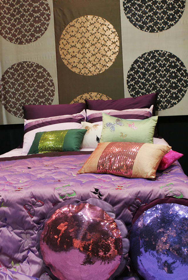 Colorido y divertido dormitorio. Cama con colcha violeta y mariposas bordadas de colores, cojines de distintas formas y colores, redondos con lentejuelas, rectangulares con aplicaciones de lentejuelas. En el muro como respaldo una tela con motivos circulares en café, dorado y negro.