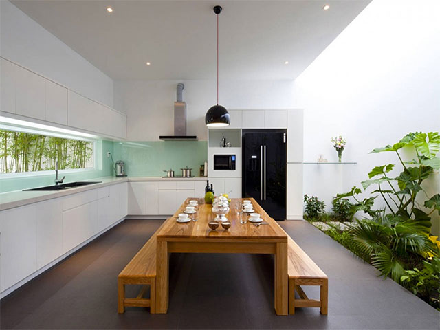 Cocina y comedor incluido con decoración zen. Paredes blancas, línea de ventanal horizontal sobre el lavaplatos en verde agua, a través de esa ventanas se aprecia un jardin con bambú. Al costado contrario de la ventana un jardín interior con helechos, palmeras y plantas de hojas grandes. La mesa de comedor es de madera  natural con banquetas a ambos lados de la mesa.