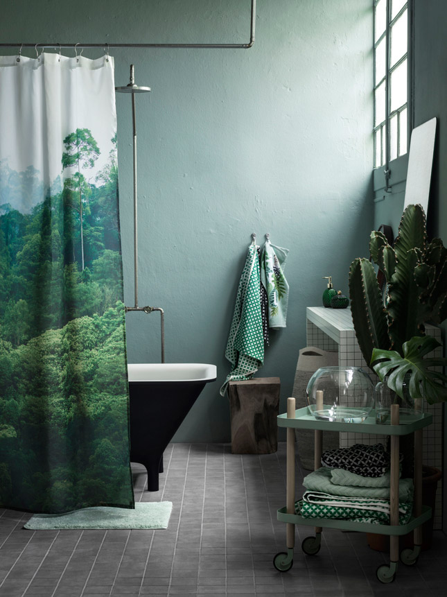 Baño en verde con un toque forest y botánico. Las paredes y accesorios como toallas y jabonera, en tonos verde botella que varían del más pálido al intenso. El toque forest lo podemos ver en un tronco usado como piso y en la gran imagen de un bosque en la cortina de baño. El look botánico lo encontramos en el cactus gigante al costado del lavamanos y en la hoja de monstera deliciosa en el frasco de vidrio que se encuentra sobre el carrito con ruedas.