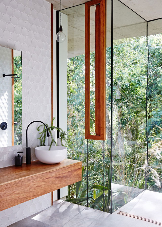 Baño con paneles vidriados con vista a un jardín muy frondoso. Inserta en los paneles, una ventana angosta con marco de madera color natural al igual que mueble donde se encuentra el vanitorio redondo de loza blanca.