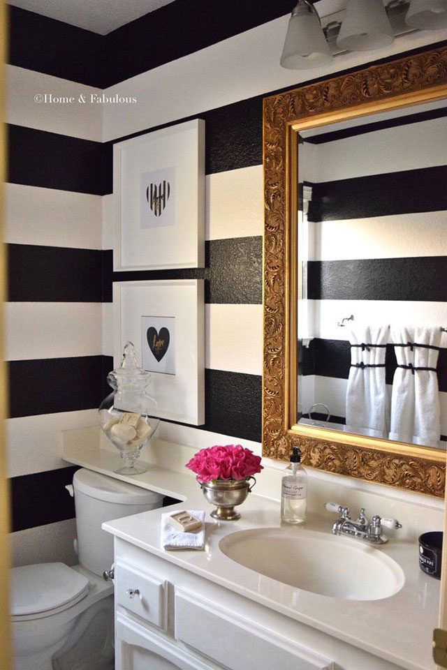 Baño con muro bicolor de líneas horizontales blancas y negras, los muebles del baño son blancos y sobre el vanitorio un espejo rectangular de marco dorado estilo rococó.