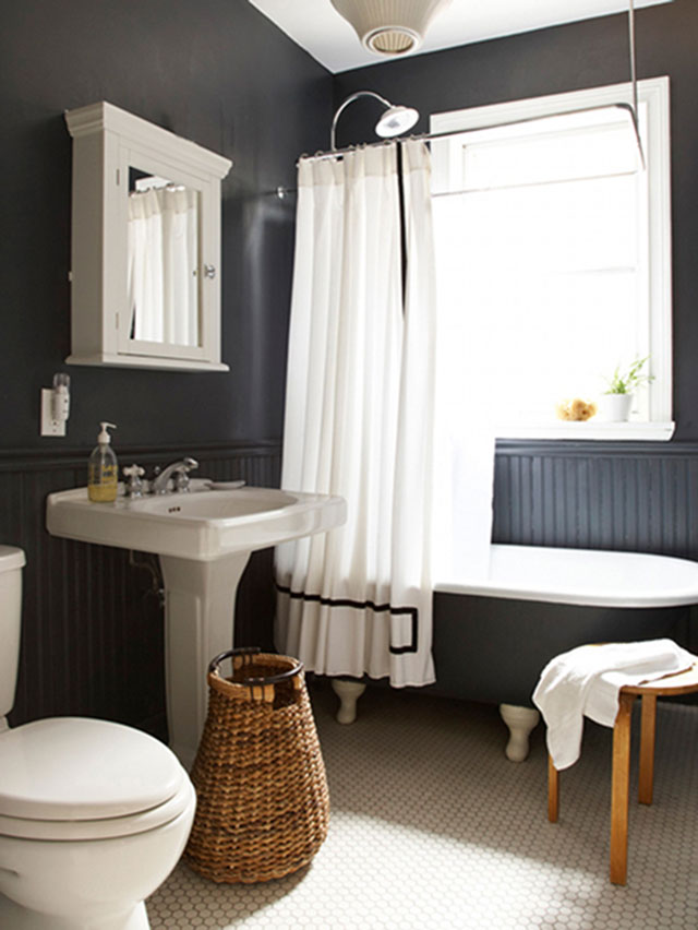 Sala de baño de paredes color gris oscuro y suelo de trama hexagonal. La tina de baño enlozada de exterior del mismo color gris oscuro de las paredes se encuentra junto a la ventana. Decoran la sala un canasto de mimbre en forma de pera y asas metálicas negras y una mesita baja redonda de madera.