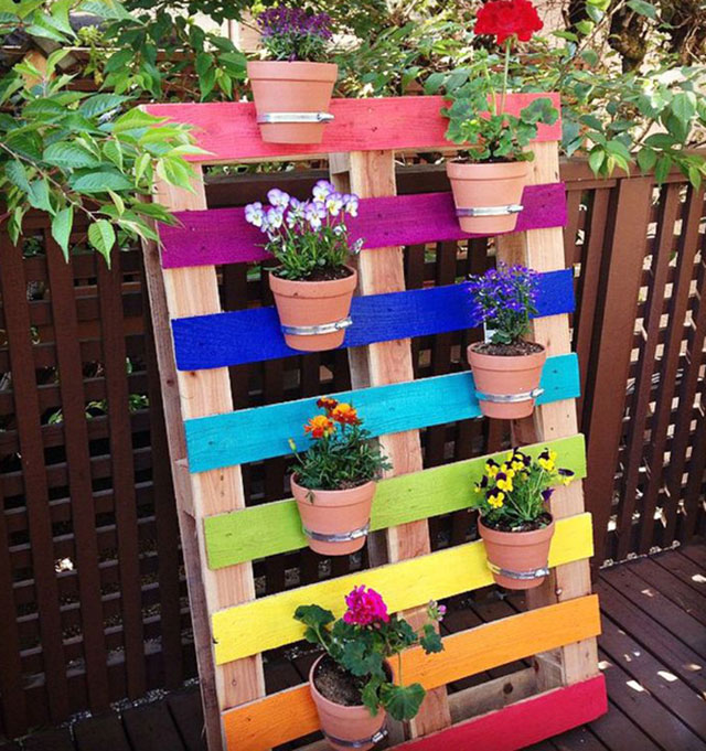 Pallet apoyado junto a una cerca con cada uno de sus travesaños pintados de color, de ellos cuelgan macetas con flores.