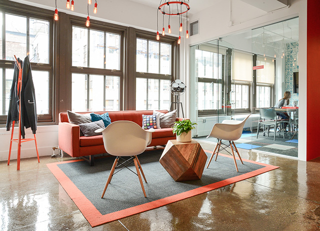 Sala de estar con sillón rojo, dos sillas y poliedro de madera como mesa de centro. A un costado, detrás de vidrio,la sala de reuniones.