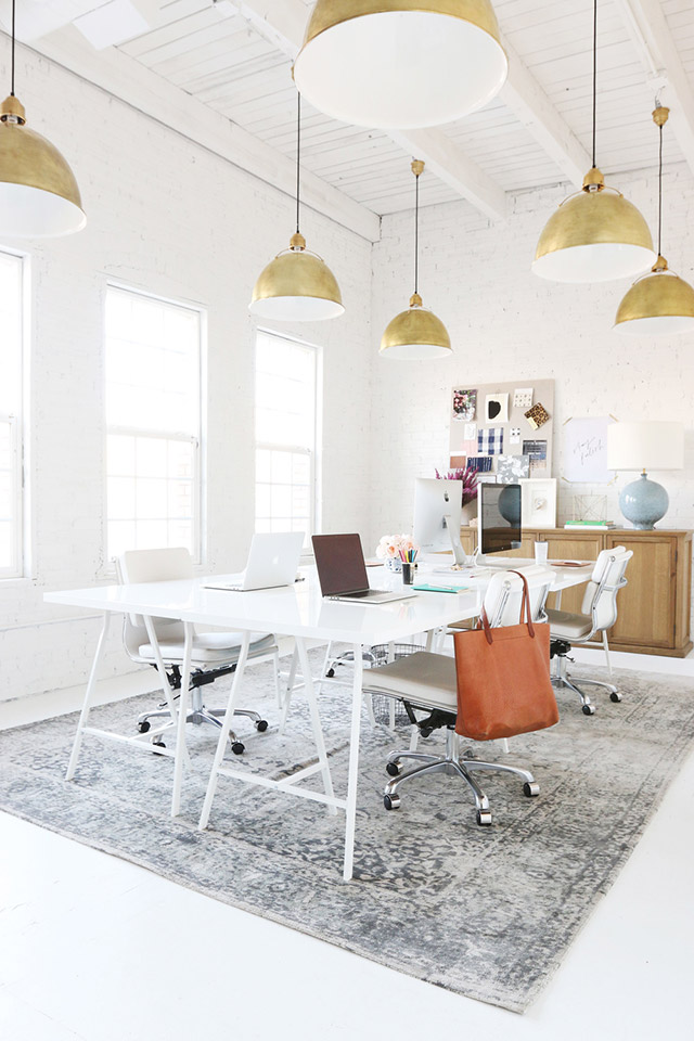 Mesa grande blanca para cuatro estaciones de trabajo. La sala es muy iluminada, de techo alto con estructura de madera a la vista y muros de ladrillos, ambos pintados blancos. Del techo cuelgan seis lámparas de estilo industrial con pantallas metálicasa doradas.